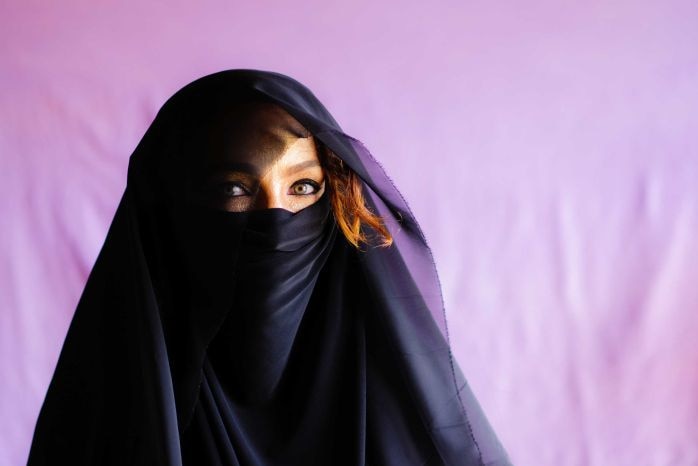 A woman covered in a black scarf with only her eyes showing looks out to the camera against a purple backdrop.