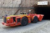 Red mining truck enters underground mine with sign reading "Big Bell Decline" above entrance.