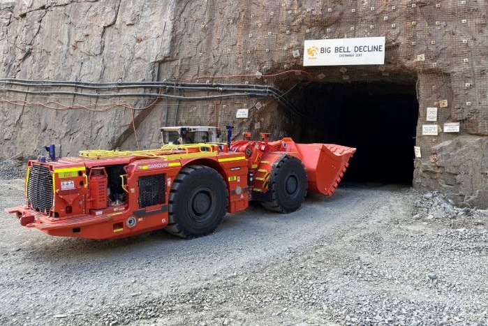 Red mining truck enters underground mine with sign reading "Big Bell Decline" above entrance.
