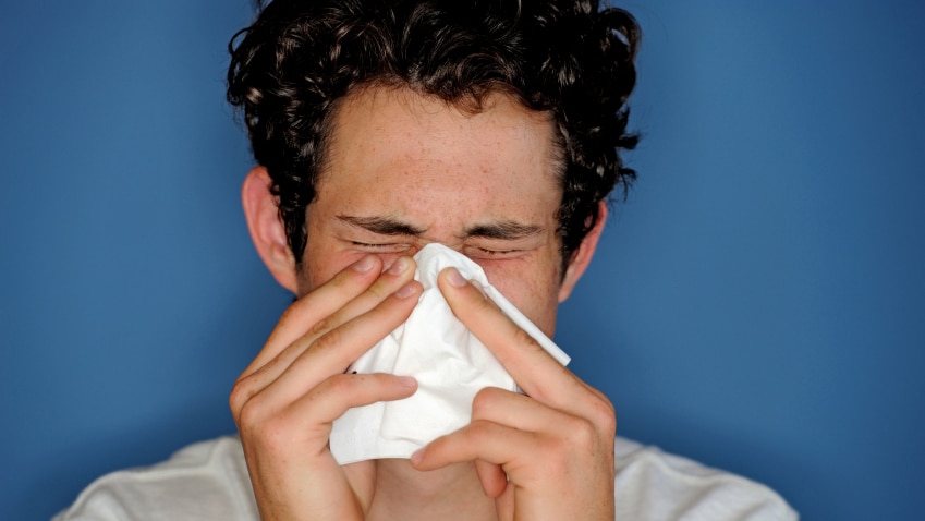 A young, dark-haired man sneezes into a tissue.