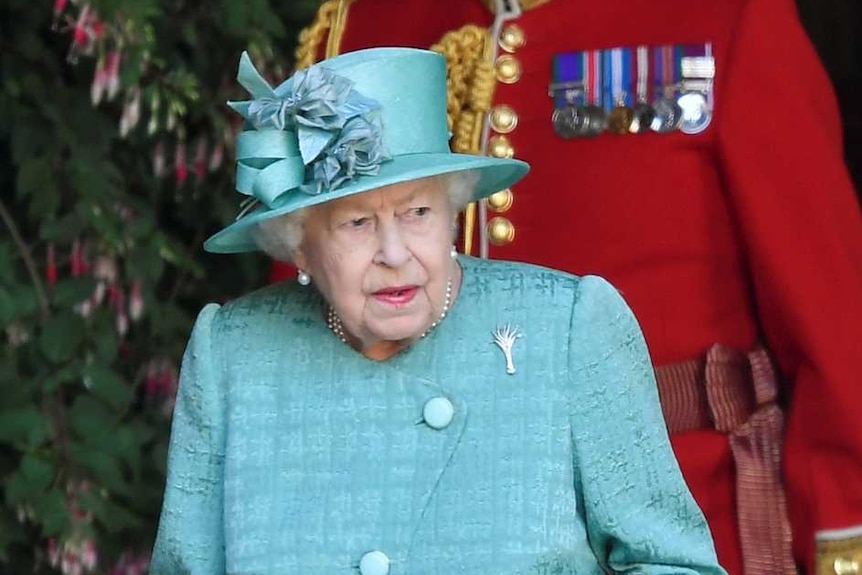 The Queen wears a blue suit and looks slightly to the right as she walks with two men in military garb, one in red, one in black