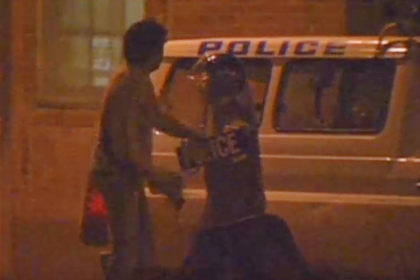 A dark still image of two people in front of a police van, taken from a video.