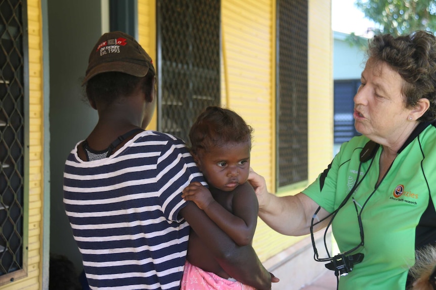 A clinic nurse examines the ear of a baby being held by her mother outside a house.