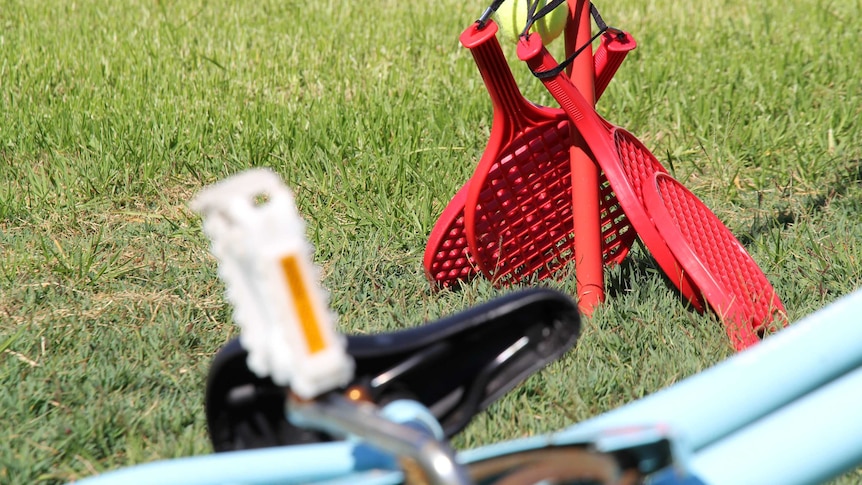 A blue bike lies abandoned on grass with a set of red children's tennis racquets behind it.
