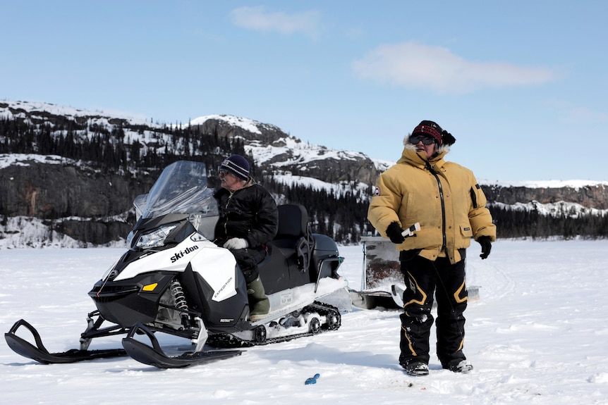 A man sits on a snowmobile, while a woman wearing a thick coat is standing next to him. 