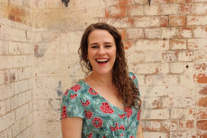 A 30-something brown-haired woman laughs. She is standing in front of an exposed brick wall and wearing a blue floral dress.