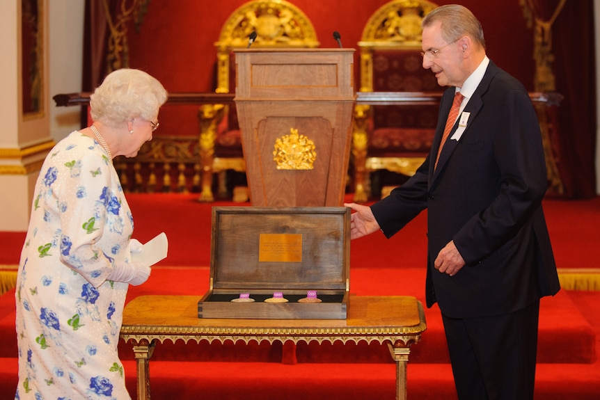 Queen Elizabeth II and Jacques Rogges stand either side of a plaque, while standing on red carpet