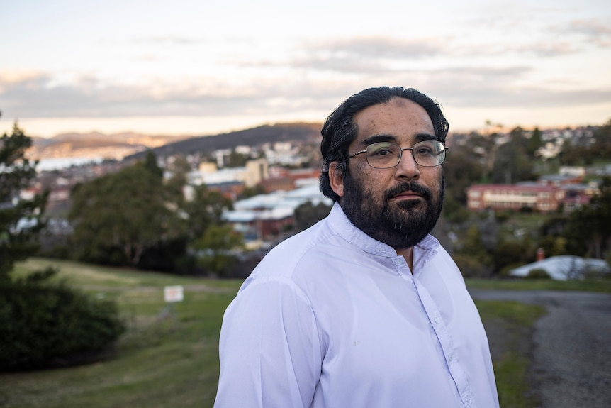 A bearded man wearing glasses stands atop a hill with houses behind him.