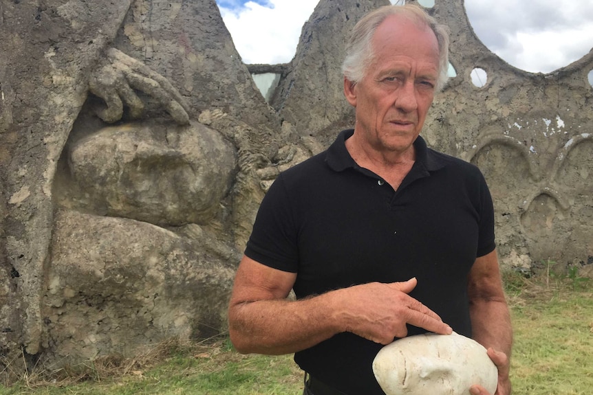 Artist Tony Phantastes holds the plaster cast mask of his son in front of his work Dreamers Gate in Collector.