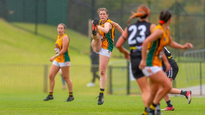 A young woman follows through after kicking a football during an Aussie rules match, with other players nearby.