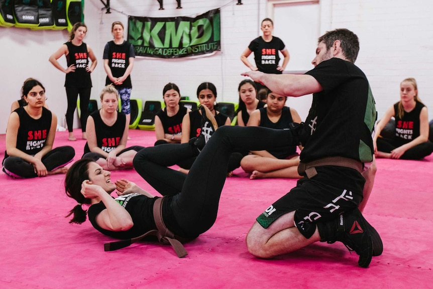 A woman and a man demonstrate a move at a self-defence class while other women sit in a circle and watch.