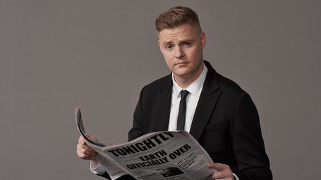 Comedian Tom Ballard reading a news paper looking pensive