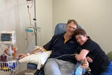 A woman hugs her husband who is hooked up to an IV in hospital.  