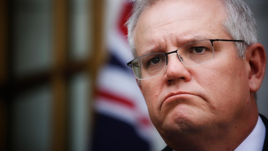 Close up of Morrison with a frown, out-of-focus Australian flag in background.