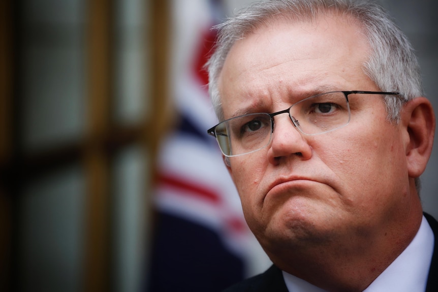 Close up of Morrison with a frown, out-of-focus Australian flag in background.