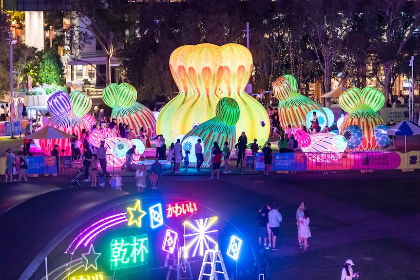 A series of large, colourful and brightly light inflatable sculptures with little faces line a walkway in a night-time garden.