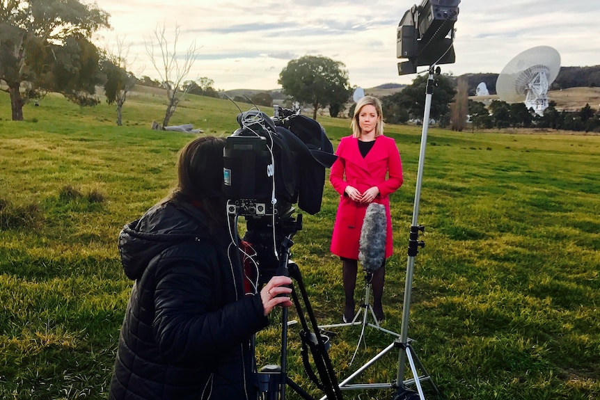 Woman operating camera pointed at journalist standing in front of satellite dishes in green field.