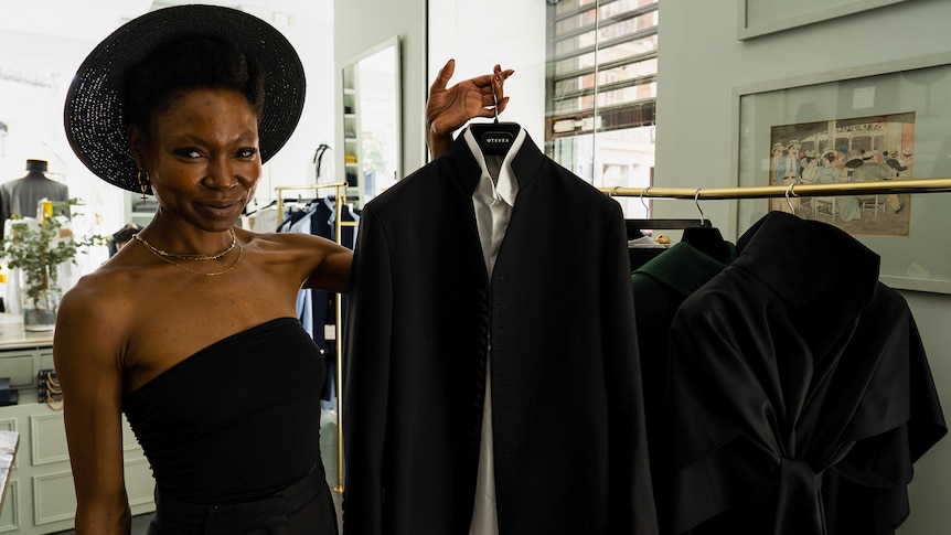 Una mujer negra con sombrero y vestido negros tiene una camisa blanca y un blazer negro.