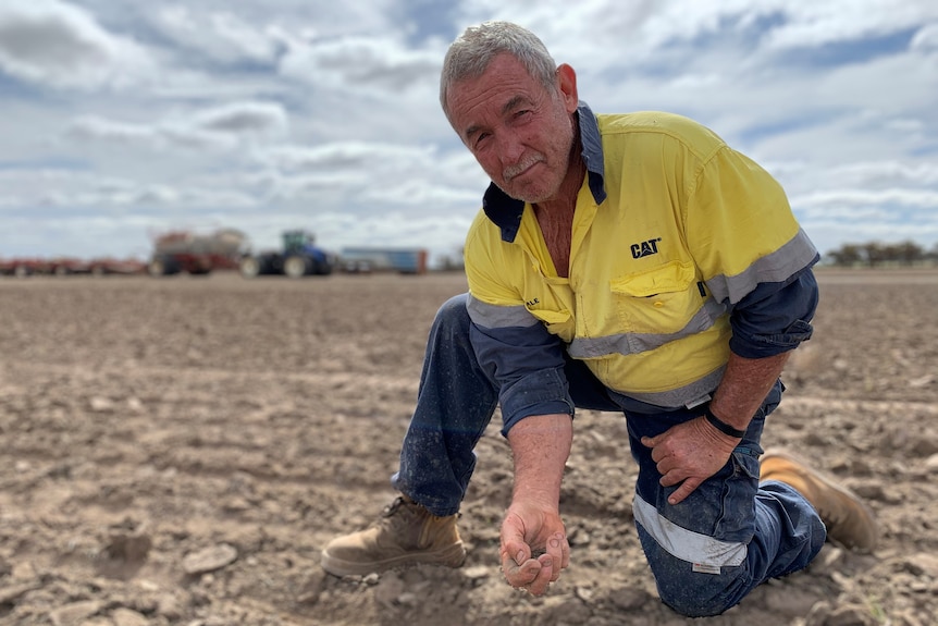 A middle-aged man wearing high visibility clothes, kneeling down in a paddock holding soil.