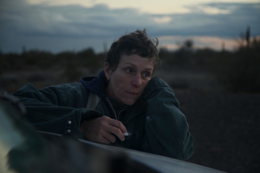 Twilight shot of the actress with pixie haircut and no make-up, wearing parka and hoodie, leaning on truck bonnet and smoking.