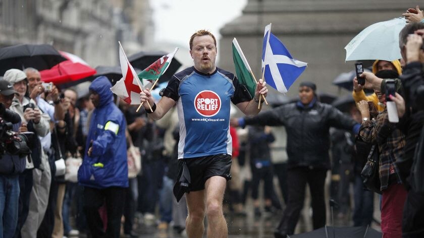 Eddie Izzard runs in a marathon while holding flags from various countries.