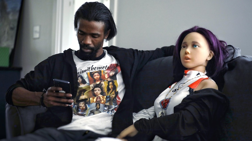 A man sits on a couch looking at his phone, a life-size doll with purple hair is next to him. 