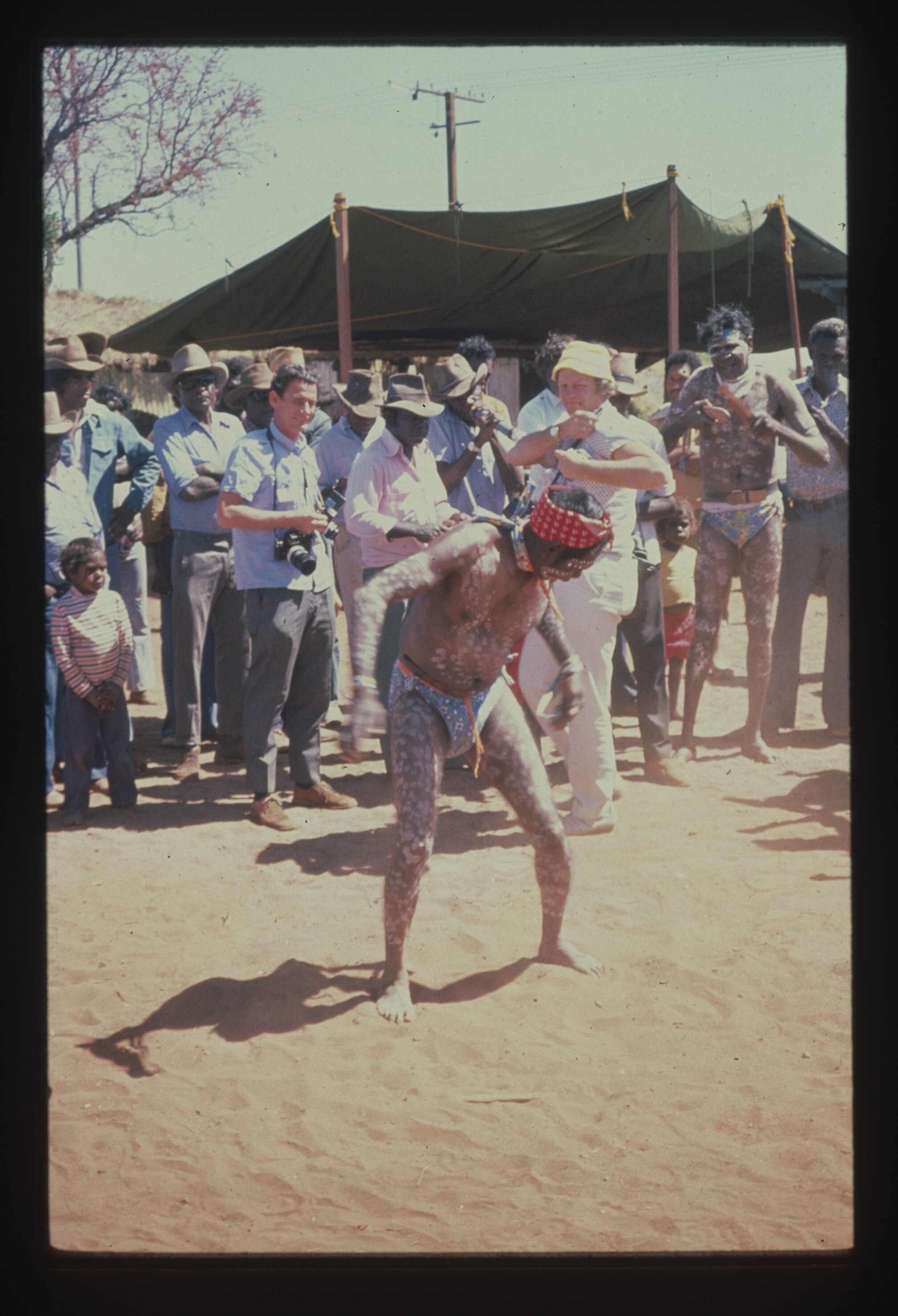 An Aboriginal man performing a cultural dance, paint on skin, red bandana on head. Crowd of photographers watching.