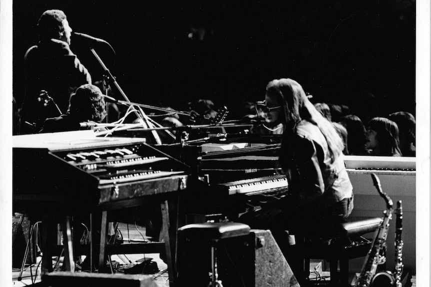 On stage, a middle-aged man sings at a microphone, another sits behind a piano and keyboards, while another sits with his guitar