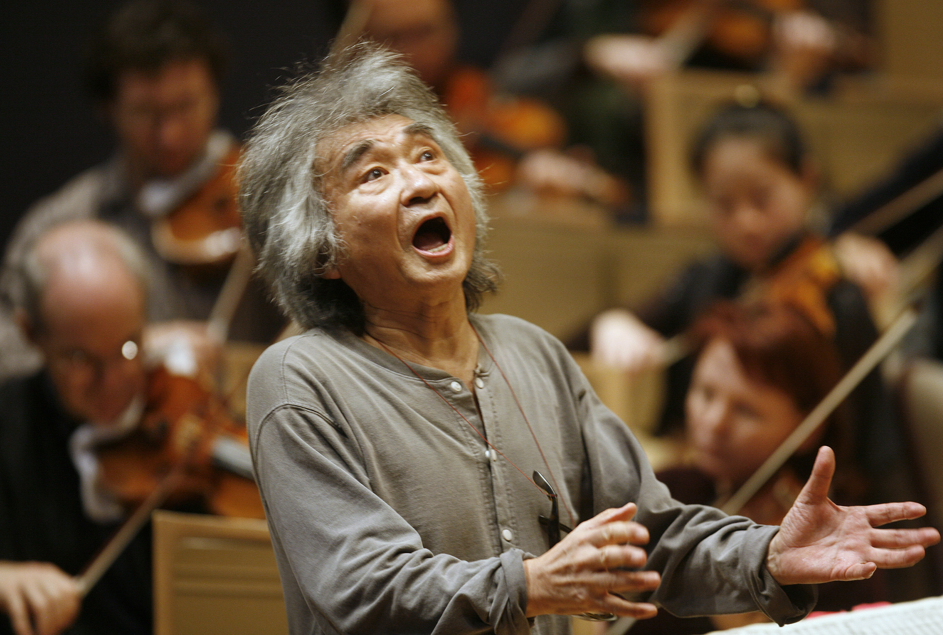 An old man in a grey shirt with his mouth open raising his hands to conduct an orchestra 