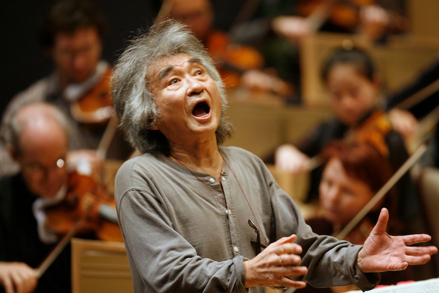 An old man in a grey shirt with his mouth open raising his hands to conduct an orchestra 