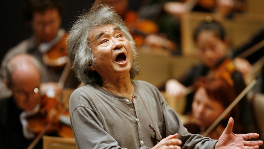 An old man in a grey shirt with his mouth open raising his hands to conduct an orchestra 