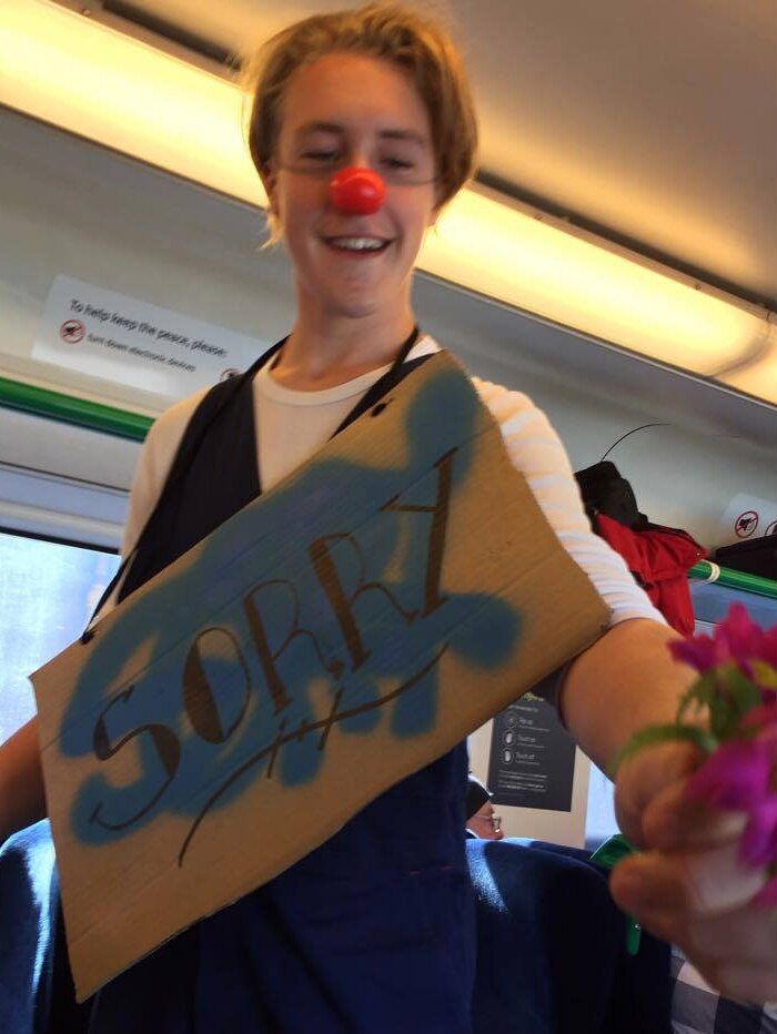 A young man wears a clown nose and a sign saying sorry while handing flowers to train passengers