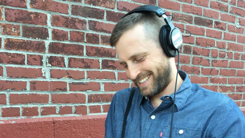 Luke Malone stands by a brick wall wearing headphones and holding a microphone and digital recorder on which he adjusts a knob.