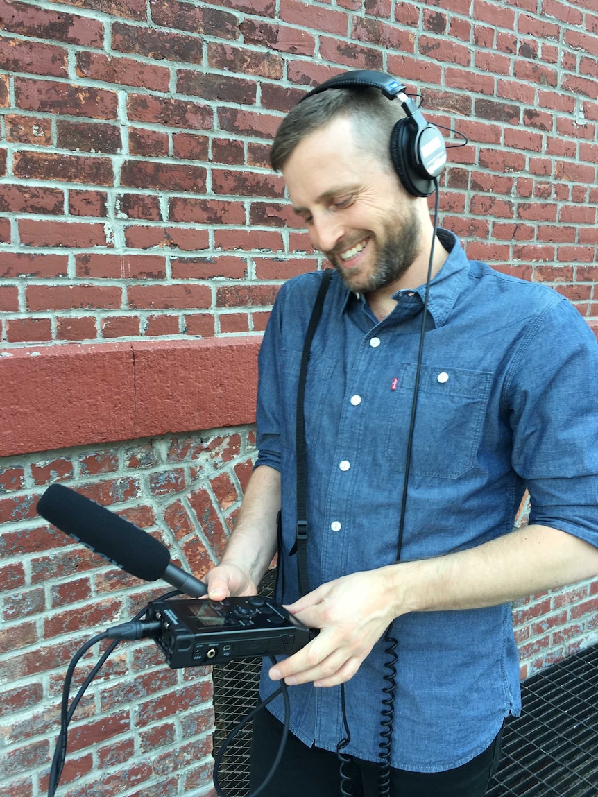 Luke Malone stands by a brick wall wearing headphones and holding a microphone and digital recorder on which he adjusts a knob.