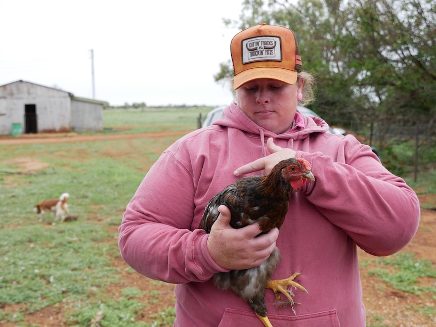 A woman wearing a hat and a hoodie stands in a paddock, stroking a chicken.