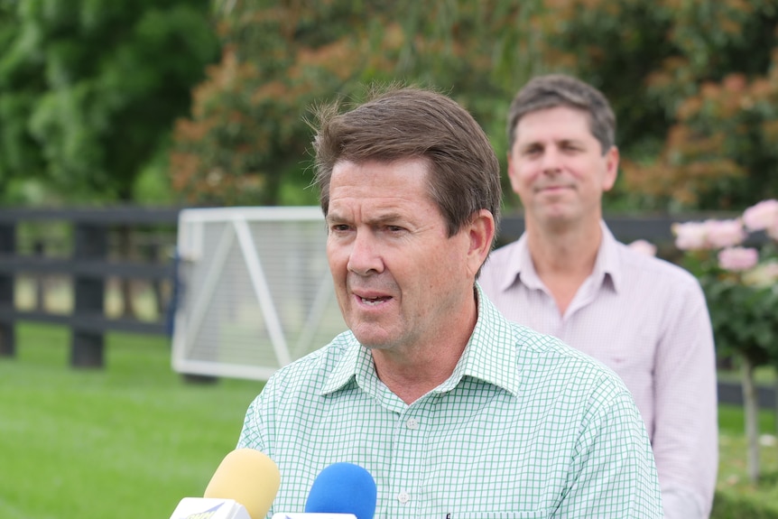 A man standing in a lush green paddock speaking in front of two microphones with another man standing in the background
