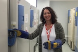 A woman wearing large blue gloves smiles as she opens one of a row of refrigerators