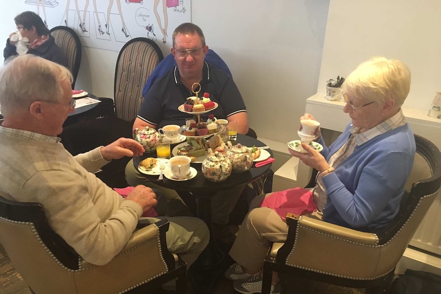Three people sit around a table with tea pots and a platter of cakes.