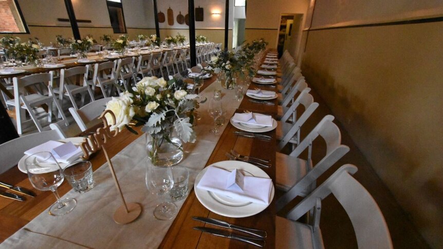 Two long tables run through a room at a wedding venue, with white roses, plates and cutlery set up on top.