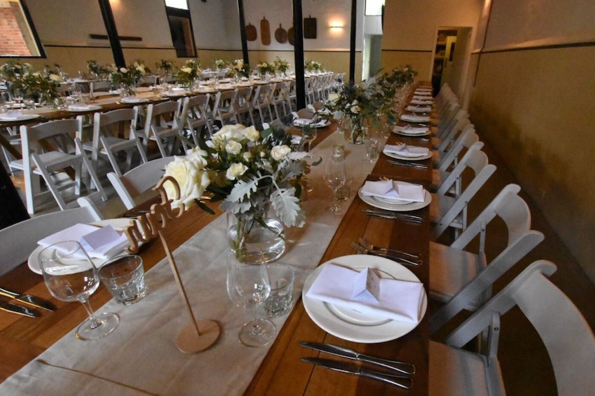 Two long tables run through a room at a wedding venue, with white roses, plates and cutlery set up on top.