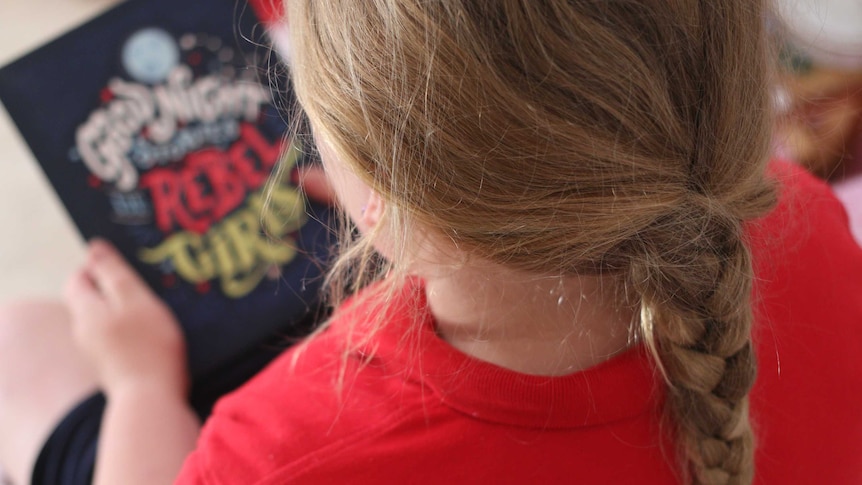 Over-the-shoulder photograph of a girl with blond hair holding an out-of-focus cover of a book.