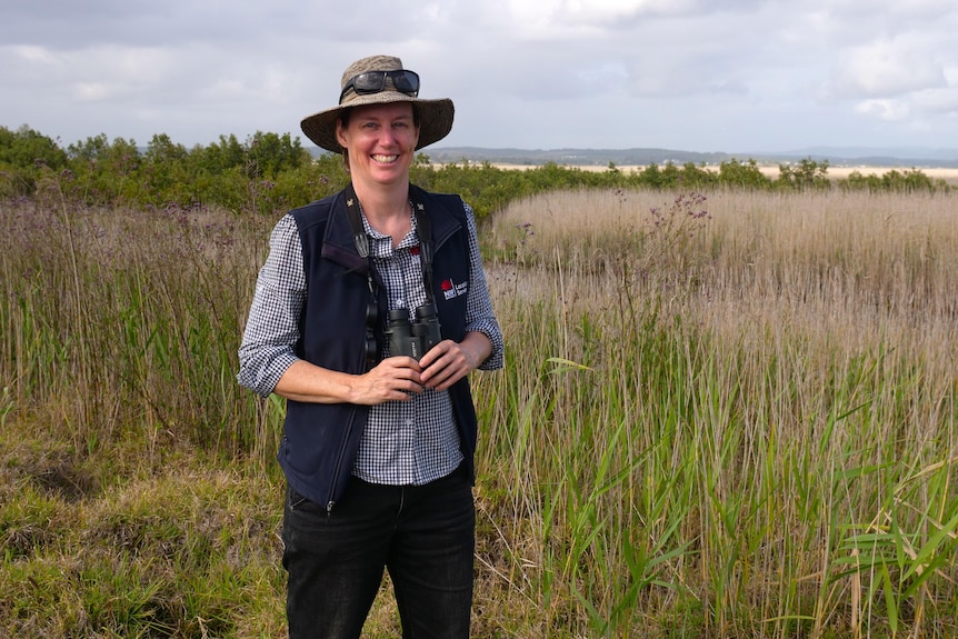 A woman wearing a hat and holding binoculars stands amoungst wetland reeds and is smiling