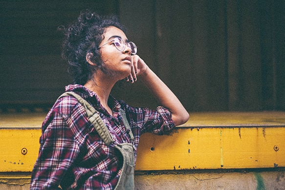 Woman in dungarees and glasses leaning on a platform and looking thoughtful.