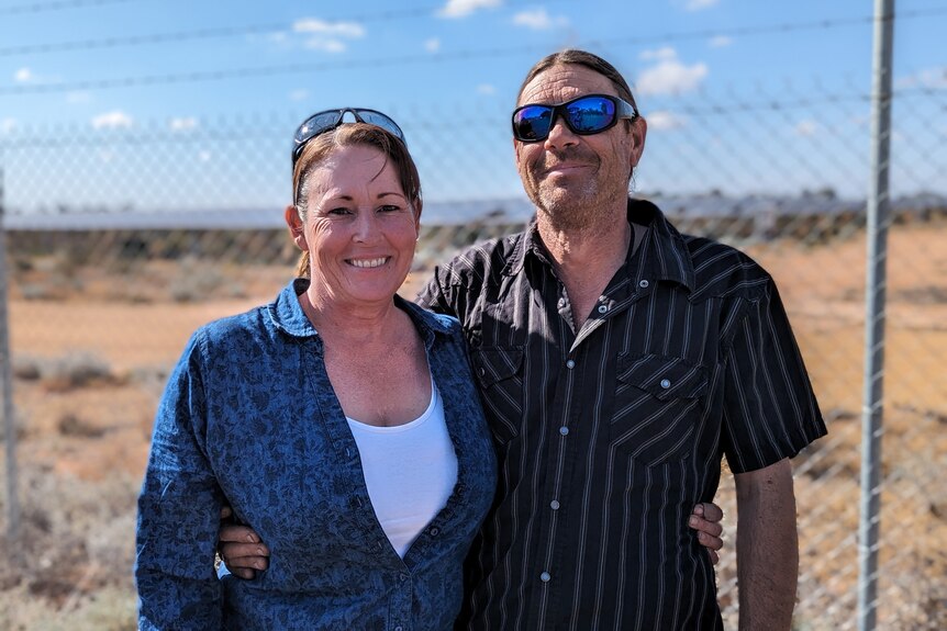 a woman and man standing together in front of a chainlink fence with dirt and scrub blurred behind them