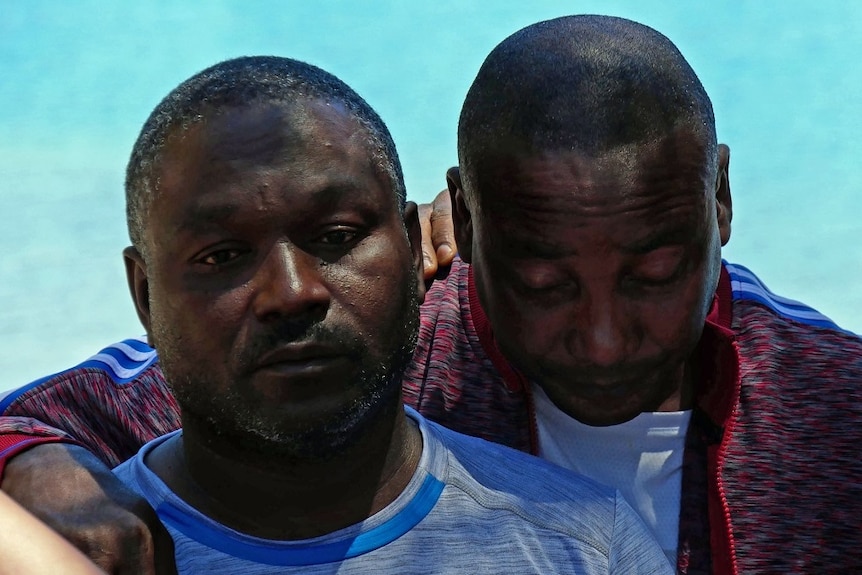 Two men console each other in front of a blue lake