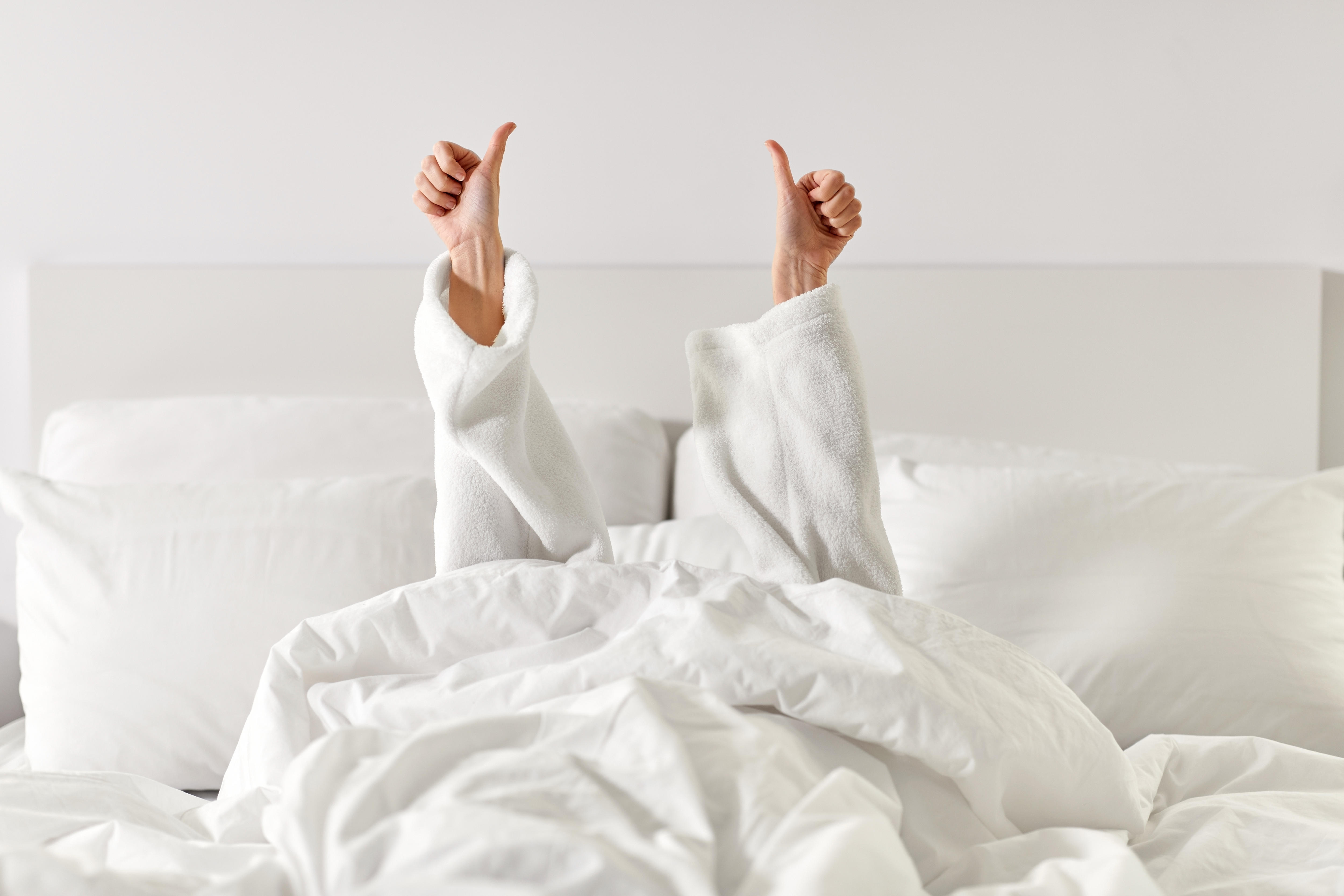 A woman's arms, in a white toweling bath robe, reach up from a hotel bed giving thumbs-up signs. 