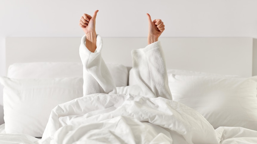 A woman's arms, in a white toweling bath robe, reach up from a hotel bed giving thumbs-up signs. 