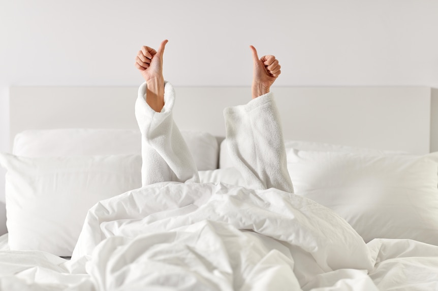 A woman's arms, in a white toweling bath robe, reach up from a hotel bed giving thumbs-up signs. 
