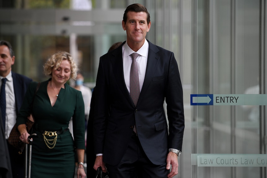 A tall man in a suit walks out of a glass door with lawyers behind him