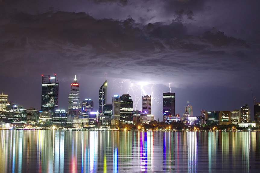 A series of bolts of lightning strike the brightly lit Perth skyline at night, with the skyline reflected in the Swan River.